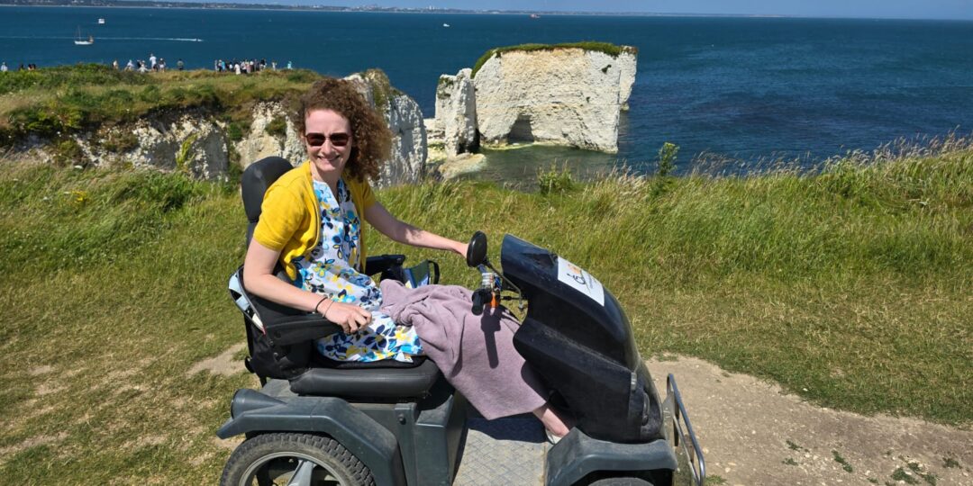 Hannah sitting on a tramper (like an off road mobility scooter) on the cliff top with Old Harry Rocks, Studland bay, in the background and the sea. It is glorious sunshine. She is wearing a white linen dress with yellow, turquoise and navy blue flowers and a yellow/mustard short cardigan. 1 of 5 wheelchair accessible things to do in Swanage.