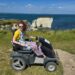 Hannah sitting on a tramper (like an off road mobility scooter) on the cliff top with Old Harry Rocks, Studland bay, in the background and the sea. It is glorious sunshine. She is wearing a white linen dress with yellow, turquoise and navy blue flowers and a yellow/mustard short cardigan. 1 of 5 wheelchair accessible things to do in Swanage.