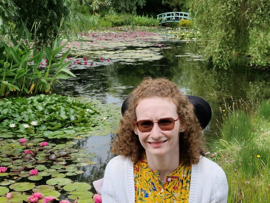 Hannah sitting in her wheelchair at Bennetts Water Garden. Behind her is a large pond with pink and white water lilies and a Monet style Japanese bridge in the distance over the bridge.