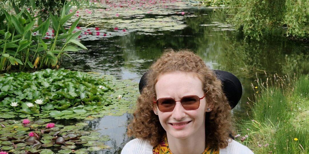 Hannah sitting in her wheelchair at Bennetts Water Garden. Behind her is a large pond with pink and white water lilies and a Monet style japanes bridge in the distance over the bridge.