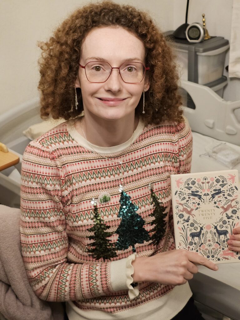 Hannah sitting down next to her bed. She is wearing a rust, pink and cream striped jumper with three sequin Christmas trees on it. She has auburn curly hair, silver earrings and large red glasses. She is smiling at the camera. She is holding up her 2025 diary which has hares, deers and foxes on it in pink and green.