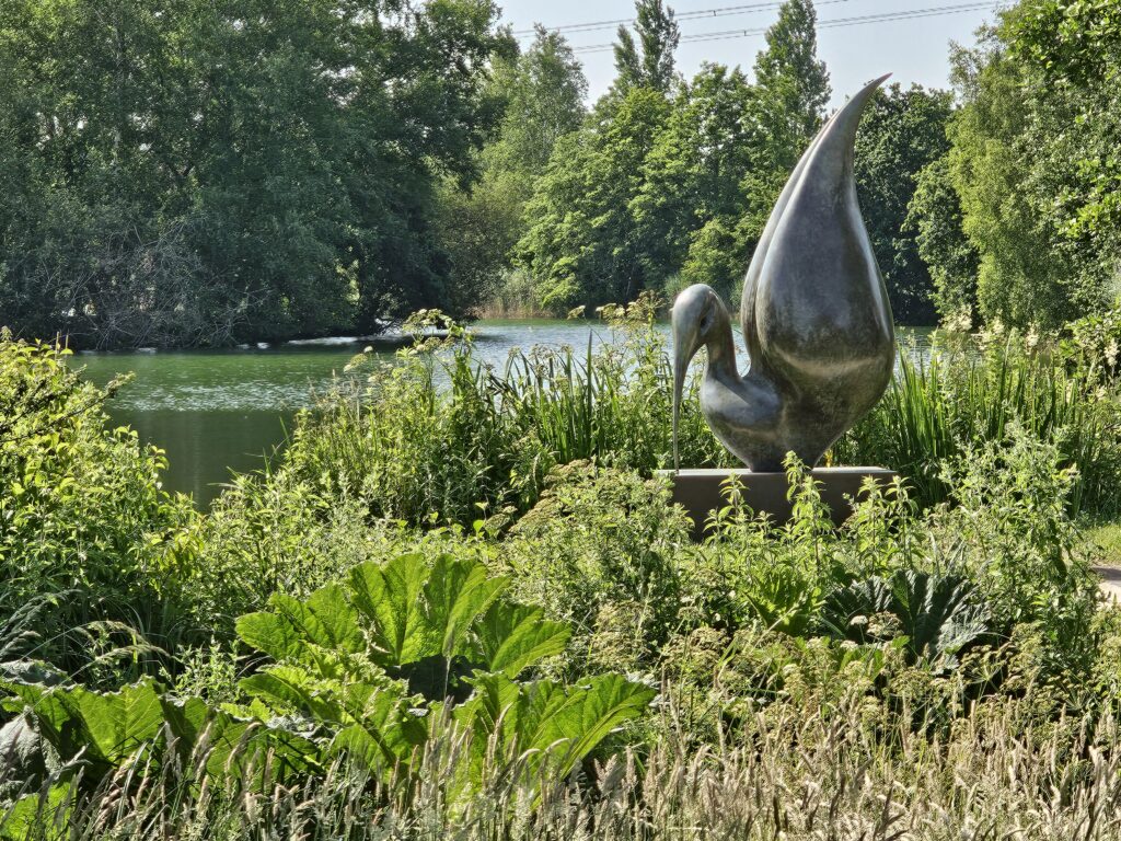 A sculpture by the lake. In front of the lake is greenery, then there is the lake with a sculpture in it and then trees behind.