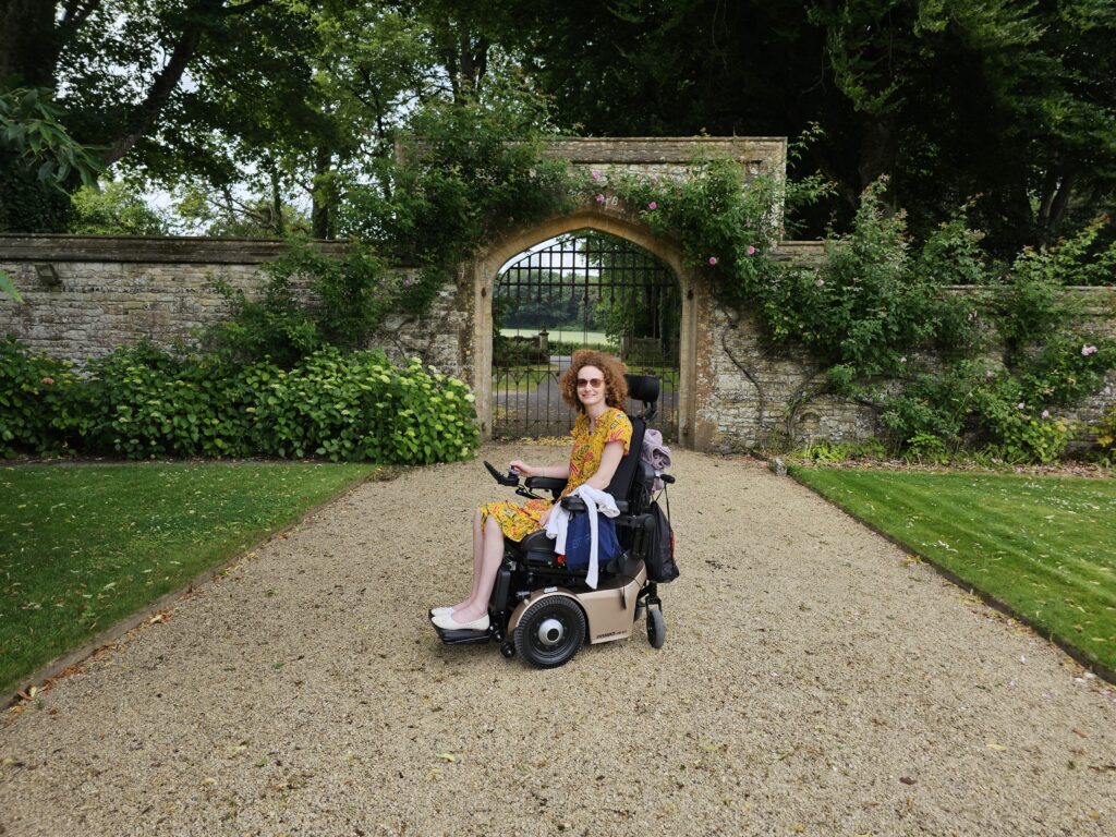Hannah by the entrance gates at Athelhampton. She is sitting in the powerchair on compact gravel, there is grass on the left and right and a brick arch entrance with gates behind her with greenery growing on it.