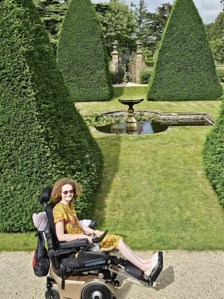 Hannah in Athelhampton gardens. There is a fountain behind her and several pyramid shaped sculptured trees.