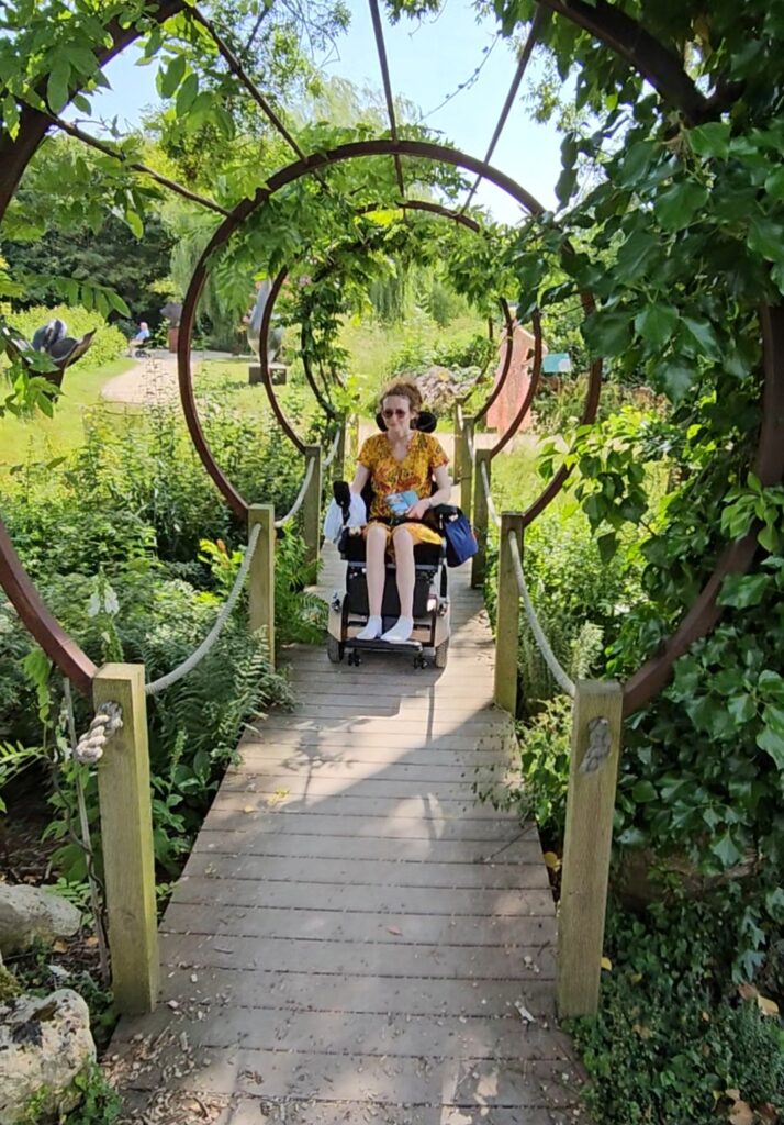 Hannah in the Sculpture by the Lakes on a trip out. She is going over a wooden bridge in her powerchair. For illustrative purposes of a trip out you could take a disabled person to.
