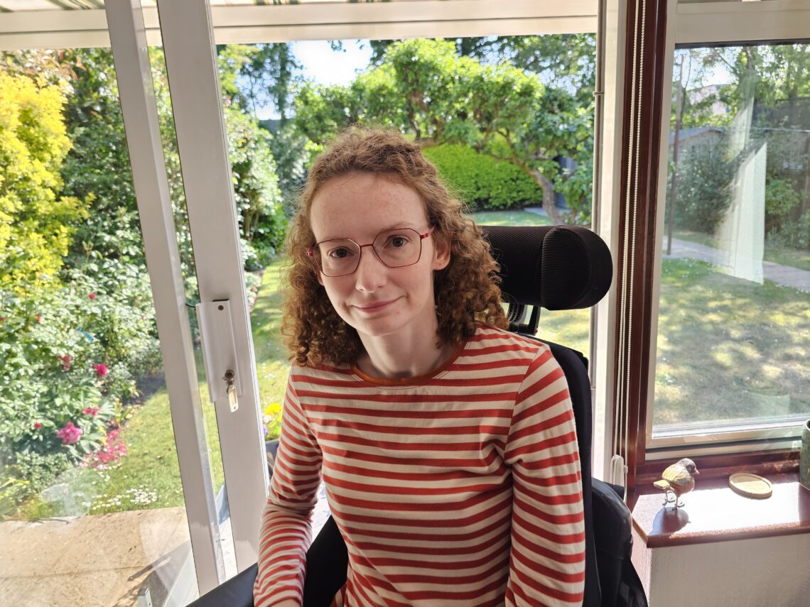 Hannah, a white woman with shoulder length auburn curly hair and red glasses, sitting in her powerchair. She is wearing an orange striped long-sleeved t-shirt and looking defiantly at the camera. To illustrate ' I use a wheelchair but I am not incapable'.